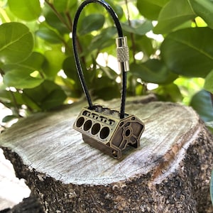 May include: A bronze-colored miniature engine block keychain. The keychain features a detailed replica of an engine block with multiple cylinders. It has a black cord and a silver-colored clasp. The keychain is displayed on a tree stump against a backdrop of green foliage.