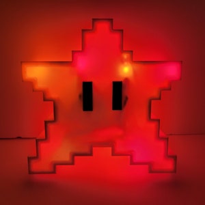 May include: A red pixelated star-shaped night light with two black eyes. The light is on and glowing.
