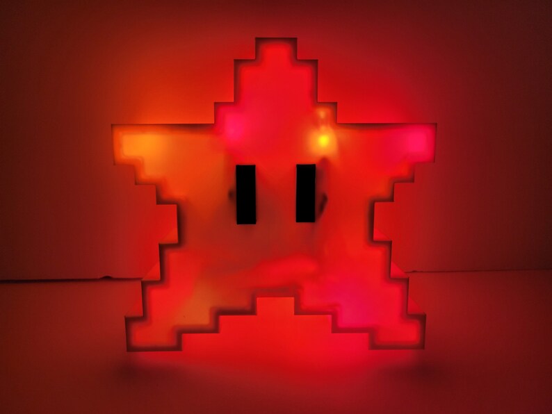 May include: A red pixelated star-shaped night light with two black eyes. The light is on and glowing.