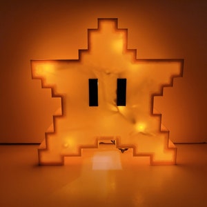 May include: A yellow pixelated star-shaped night light with a face. The light is on and glowing.