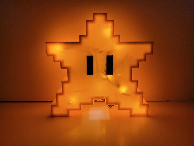 May include: A yellow pixelated star-shaped night light with a face. The light is on and glowing.
