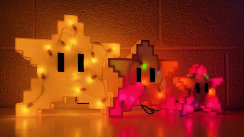May include: Three pixelated star-shaped light-up figures. The largest is yellow with white lights, the middle is pink with red lights, and the smallest is red with orange lights.