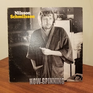 May include: A black and white vinyl record album cover featuring a man in a striped robe with the text "Nilsson Schmilsson" and "NOW-SPINNING" printed on the front.