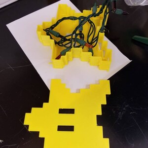 May include: A yellow plastic star-shaped mold with a set of green Christmas lights inside. The lights are strung through the mold and have small, clear bulbs. The mold is sitting on a white piece of paper.