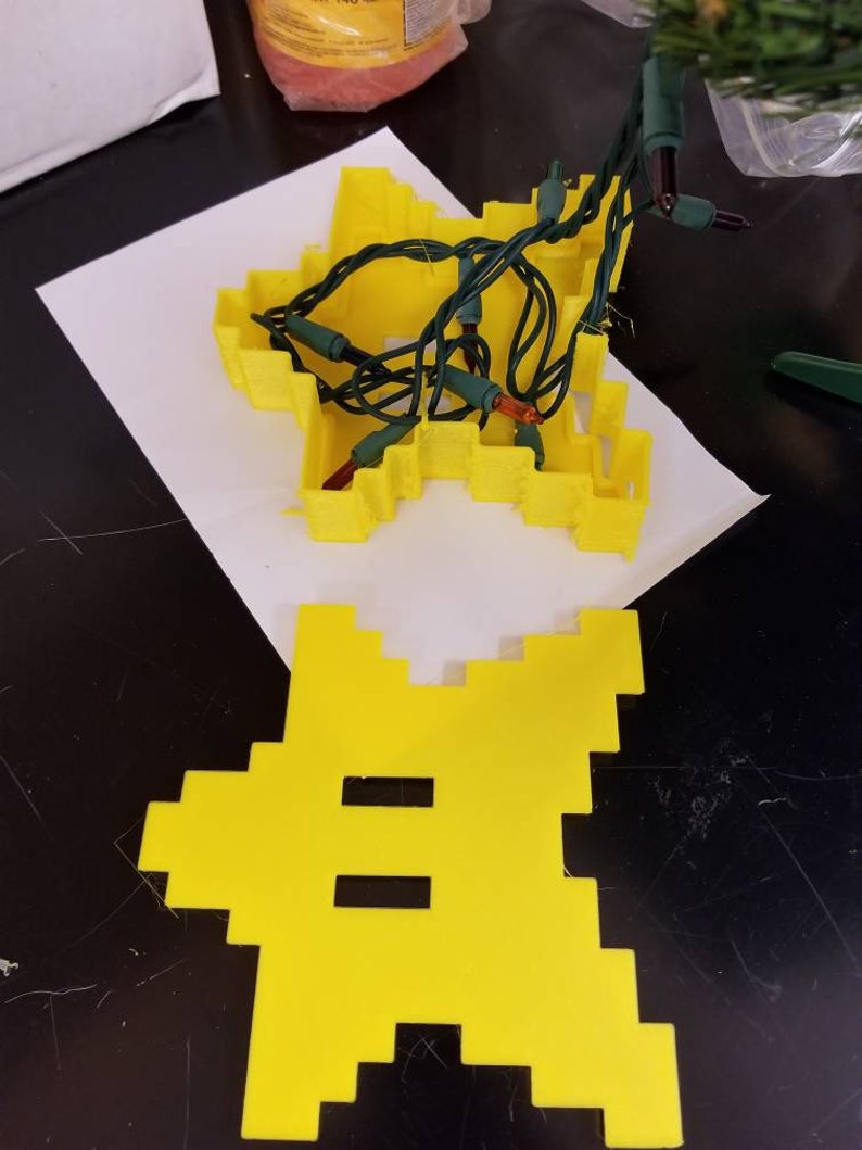 May include: A yellow plastic star-shaped mold with a set of green Christmas lights inside. The lights are strung through the mold and have small, clear bulbs. The mold is sitting on a white piece of paper.