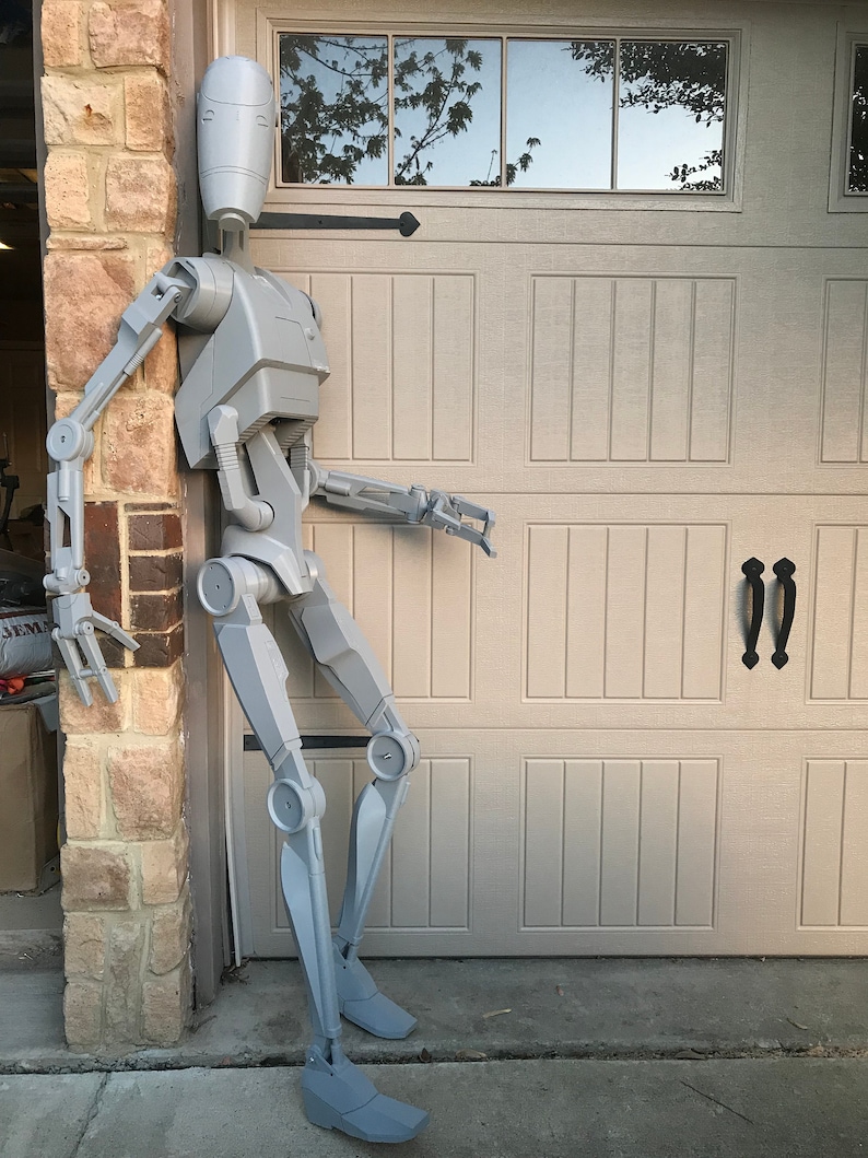 Może przedstawiać: Szara, członkowa figurka robota stojąca obok garażowej bramy. Robot ma ludzką sylwetkę z dużą głową i długimi kończynami.
