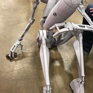 Może przedstawiać: Szary, artykułowany model robota o ludzkim kształcie. Robot jest w pozycji siedzącej z wyciągniętymi ramionami i zgiętymi nogami. Robot ma szczeg&oacute;łowy projekt z widocznymi połączeniami i przekładniami.