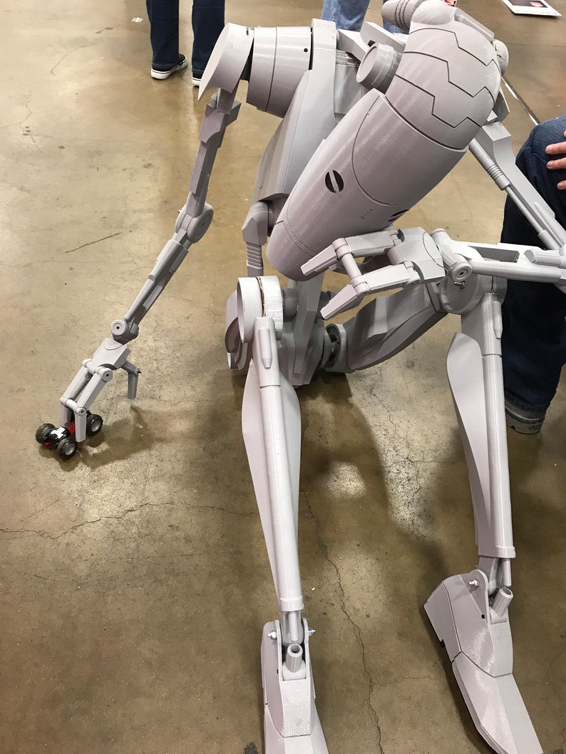 Może przedstawiać: Szary, artykułowany model robota o ludzkim kształcie. Robot jest w pozycji siedzącej z wyciągniętymi ramionami i zgiętymi nogami. Robot ma szczeg&oacute;łowy projekt z widocznymi połączeniami i przekładniami.