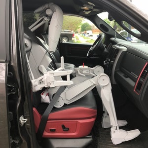 Może przedstawiać: Szara figurka robota siedzi na miejscu kierowcy czarnego pickupa. Robot ma zapięty pas bezpieczeństwa i wyciągnięte ręce. Wnętrze pickupa jest czerwone i czarne.