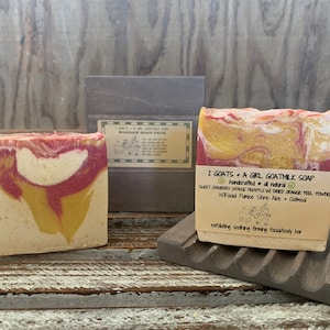 以下が含まれることがあります： 赤、黄、白の渦巻き模様が入った手作りヤギミルク石鹸が2つ。石鹸には「2 Goats + A Girl Goatmilk Soap」とラベルが貼られており、成分として「Sweet Cranberry Orange Pineapple w/ Dried Orange Peel Powder, W/Facial Pumice Stone Aloe + Oatmeal」が記載されています。ラベルには「Exfoliating Soothing Firming Facial/Body Bar」とも書かれています。