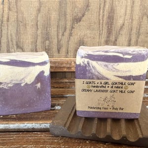 Puede incluir: Dos barras de jabón artesanal de leche de cabra con aroma cremoso a lavanda. El jabón es morado con remolinos blancos y está etiquetado como "2 GOATS + A GIRL GOATMILK SOAP". El jabón está sobre una jabonera marrón.