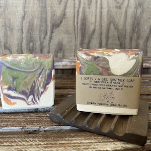 Puede incluir: Dos barras de jabón de leche de cabra hecho a mano con un remolino de colores naranja, verde y morado. El jabón está etiquetado como "2 Goats A Girl Goatmilk Soap" y "Pineapple Mango Papaya Exfoliating Goat Milk Bar with Added Kale Acid, Vitamin C, Vitamin E3. Exfoliating Moisturizing Vitamin Rich Bar."
