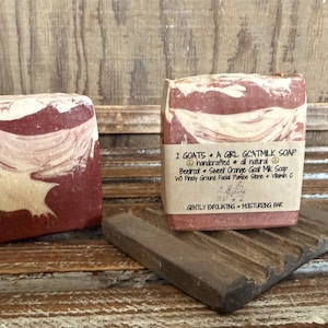 May include: Two rectangular bars of handmade goat milk soap with a marbled red and cream design. One bar rests on a wooden soap dish. The soap is labeled "2 GOATS + A GIRL GOATMILK SOAP" with ingredients listed. The soap is a gently exfoliating and moisturizing bar.