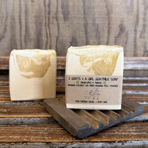 Puede incluir: Dos barras de jabón de leche de cabra de color beige claro con un diseño veteado. El jabón está etiquetado "2 GOATS + A GIRL GOATMILK SOAP" con los ingredientes enumerados. Las barras descansan sobre un jabonera marrón oscuro.
