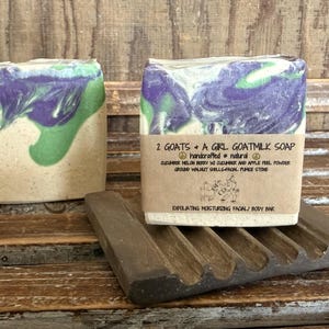 May include: Two bars of goat milk soap with purple, green, and tan swirls, one resting on a wooden soap dish. The soap is labeled "2 GOATS + A GIRL GOATMILK SOAP" and lists ingredients. The soap dish is dark brown and has grooves.