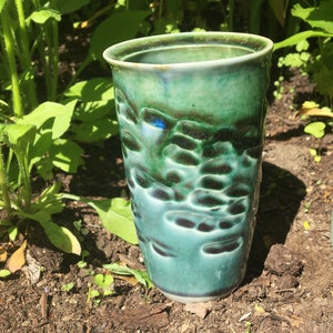 Könnte beinhalten: Eine grüne Keramiktasse mit strukturierter Oberfläche. Die Tasse hat eine dunkelgrüne Glasur mit einem hellgrünen und blauen irisierenden Schimmer.