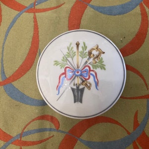 May include: A white porcelain lid with a painted design of a bow, arrows, and a key. The design is in shades of blue, red, green, and gold.