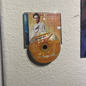May include: A gold compact disc with the text "Marc Anthony Todo A Su Tiempo" and a picture of Marc Anthony on the front. The disc is in a clear plastic case with a white background and the text "Marc Anthony Todo A Su Tiempo" on the front. The disc is signed "Evelyn".