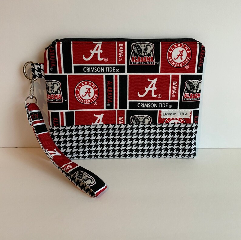 May include: A black and red zippered pouch with a black and white houndstooth pattern on the bottom. The pouch has a black strap and features a repeated pattern of the Alabama Crimson Tide logo, including the letter "A" and the words "Crimson Tide". The text "Dream Big!" is printed on the pouch.