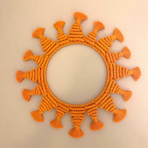 May include: A circular wall hanging featuring a central open ring adorned with radiating orange macrame cord patterns. Each section consists of tightly knotted cord ending in a small, flared tassel, creating a sunburst effect around the perimeter.