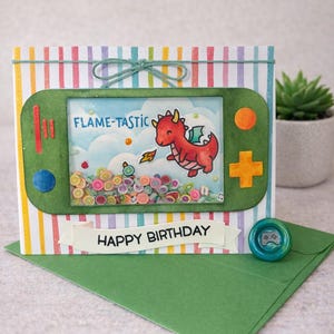 May include: A handmade birthday card with a green game console design. The card features a red dragon illustration with the text "FLAME-TASTIC" and "HAPPY BIRTHDAY". The card is decorated with colorful confetti and a green envelope.