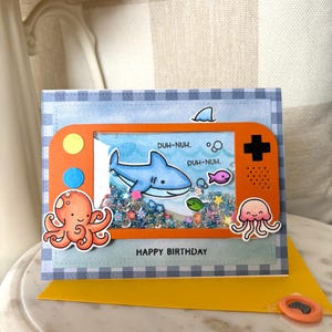 May include: A handmade birthday card with an orange game console design. The card features a shark, fish, and octopus illustrations. The text "HAPPY BIRTHDAY" is printed at the bottom. The background is blue and white checkered.