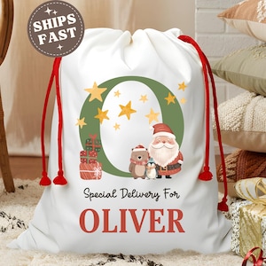 May include: White Christmas gift bag with red drawstring closure. The bag features a festive design with Santa Claus, a bear, and a penguin, along with the text "Special Delivery For OLIVER". The bag is decorated with a green wreath and gold stars.