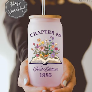 May include: A pink and white tumbler with a wooden lid and a straw. The tumbler has a design of a book with flowers coming out of it. The text on the tumbler reads "Chapter 40 First Edition 1985".