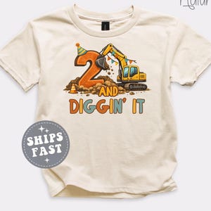 May include: A natural-colored short-sleeve t-shirt featuring a graphic of a yellow excavator digging into a large orange number two. The design includes a party hat, colorful bunting, and the text 2 AND DIGGIN' IT in orange and blue letters.