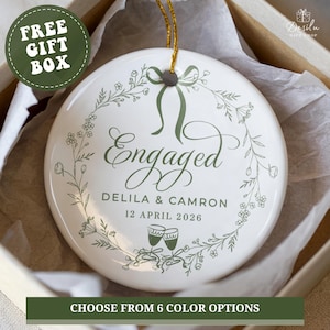 May include: A white ceramic ornament with the word "Engaged" in green script, surrounded by a floral wreath. The names Delila & Camron and the date 12 April 2026 are also printed on the ornament. The ornament is in a gift box.