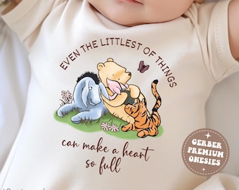 May include: A cream-colored baby onesie featuring a Winnie the Pooh design with Eeyore and Tigger. The text reads "Even the littlest of things can make a heart so full." Includes the text "GERBER PREMIUM ONESIES" and "Natural".