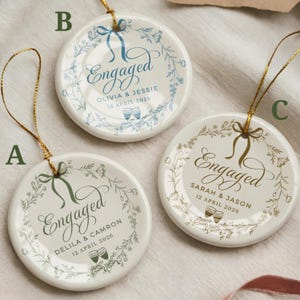May include: Three white ceramic engagement ornaments with gold string hangers. Each ornament features the word "Engaged" in elegant script, surrounded by floral designs. Names and dates are printed below, with different colour accents.
