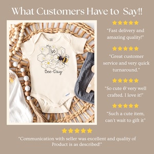 May include: Cream-colored baby onesie with a bee-themed design. The onesie features a bee, daisies, and the text "My 1st Bee-Day". The onesie is displayed on a woven mat with a wooden bead garland and a gray burp cloth.