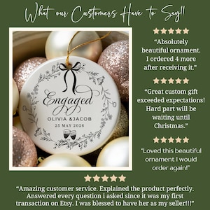 May include: A white ceramic ornament with the word "Engaged" in elegant script, surrounded by a floral wreath. The names Olivia & Jacob and the date May 25, 2026, are printed below. The ornament is hanging with a gold string, surrounded by other Christmas ornaments.