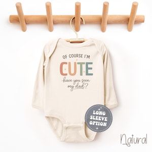 May include: A long-sleeve, natural-colored baby bodysuit with the text "OF COURSE I'M CUTE have you seen my dad?" in colorful letters. The bodysuit hangs from a wooden hanger. A round sticker says "LONG SLEEVE OPTION".