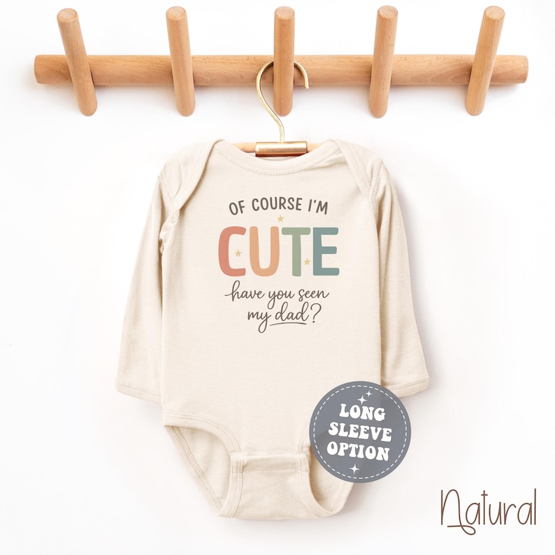 May include: A long-sleeve, natural-colored baby bodysuit with the text "OF COURSE I'M CUTE have you seen my dad?" in colorful letters. The bodysuit hangs from a wooden hanger. A round sticker says "LONG SLEEVE OPTION".