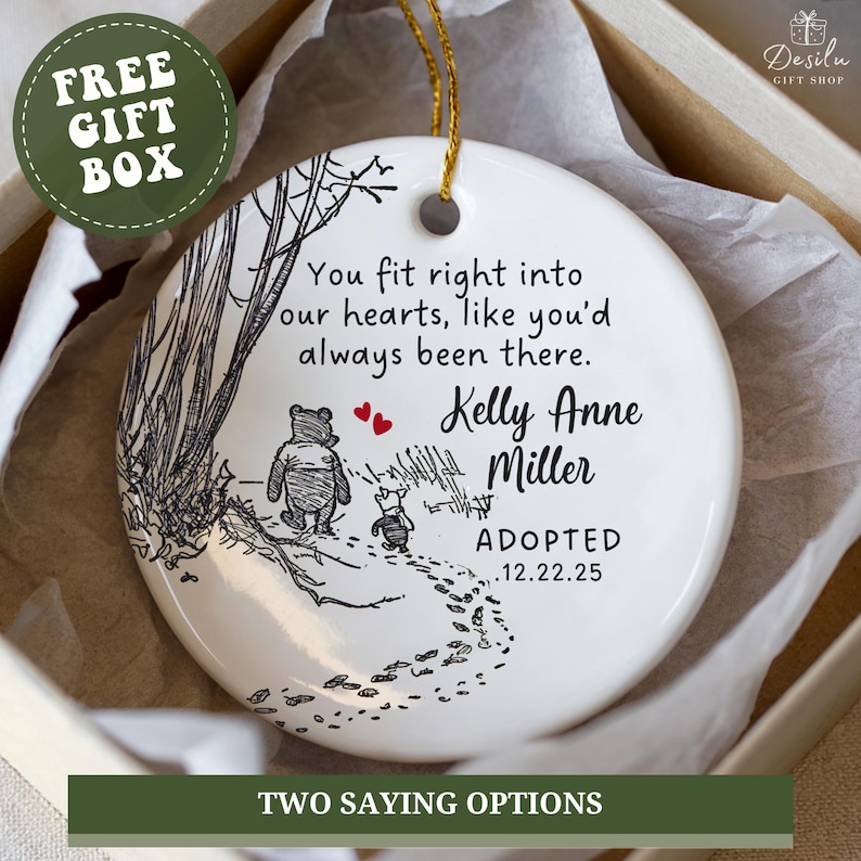 K&ouml;nnte beinhalten: Wei&szlig;er Keramik-Weihnachtsschmuck mit goldener Aufh&auml;ngeschnur. Der Schmuck zeigt eine Winnie-the-Pooh-Illustration und den Text "You fit right into our hearts, like you'd always been there." Er enth&auml;lt einen Namen und ein Adoptionsdatum.