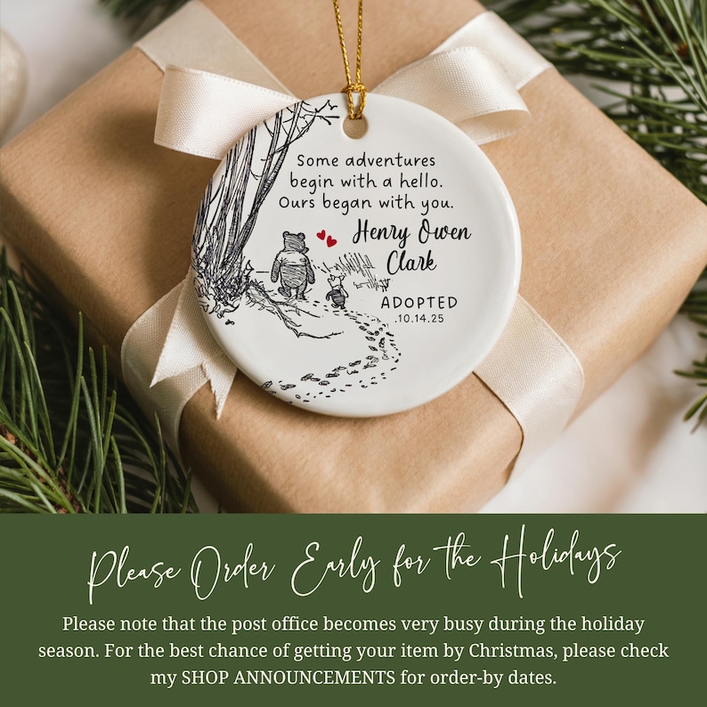 K&ouml;nnte beinhalten: Wei&szlig;er Keramikschmuck mit goldenem Band, mit einer Winnie-the-Pooh-Illustration und dem Text "Some adventures begin with a hello. Ours began with you. Henry Owen Clark ADOPTED 10.14.25". Der Schmuck liegt auf einem Geschenk.