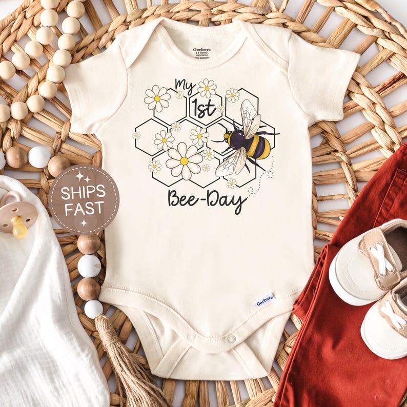 May include: Cream-colored baby onesie with a bee-themed design. The graphic includes a honeycomb, daisies, and a bumblebee, with the text "my 1st Bee-Day". The onesie is paired with red pants and white shoes.