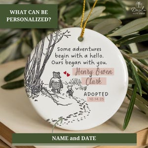 K&ouml;nnte beinhalten: Runder wei&szlig;er Keramikschmuck mit Winnie-the-Pooh-Motiv. Der Schmuck zeigt eine Skizze von Winnie Puuh und Ferkel, die auf einem Pfad gehen. Der Text enth&auml;lt "Some adventures begin with a hello. Ours began with you." und personalisierte Namen und Daten.
