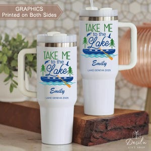 K&ouml;nnte beinhalten: Zwei wei&szlig;e Reisebecher mit dem Text "TAKE ME to the Lake" in Gr&uuml;n und Blau. Das Design umfasst ein Boot, B&auml;ume und V&ouml;gel. Der Name "Emily" und "LAKE GENEVA 2025" sind ebenfalls auf den Bechern aufgedruckt. Ein Becher hat einen Griff und der andere einen Strohhalm.