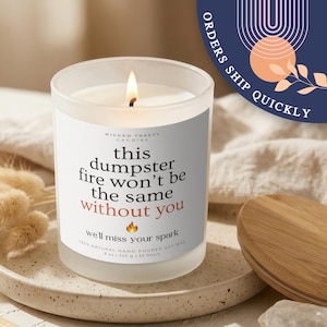 Może przedstawiać: Świeca w matowym szkle z białą etykietą zawierającą tekst: WICKED TOASTY CANDLES, this dumpster fire won't be the same without you, we'll miss your spark, 100% natural hand poured soy wax, 225 g, 35 hours.