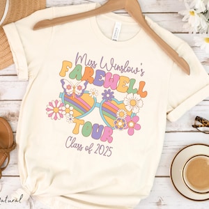 May include: A white t-shirt with a retro-style graphic design featuring a pair of sunglasses with flowers and a butterfly. The text on the shirt reads "Miss Winslow's Farewell Tour Class of 2025".