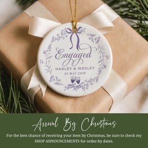 May include: A round white ceramic ornament with the word "Engaged" in purple script, encircled by a floral wreath. It features the names "Harley & Wesley" and the date "25 MAY 2026". The ornament hangs on a gold string, presented on a wrapped gift.