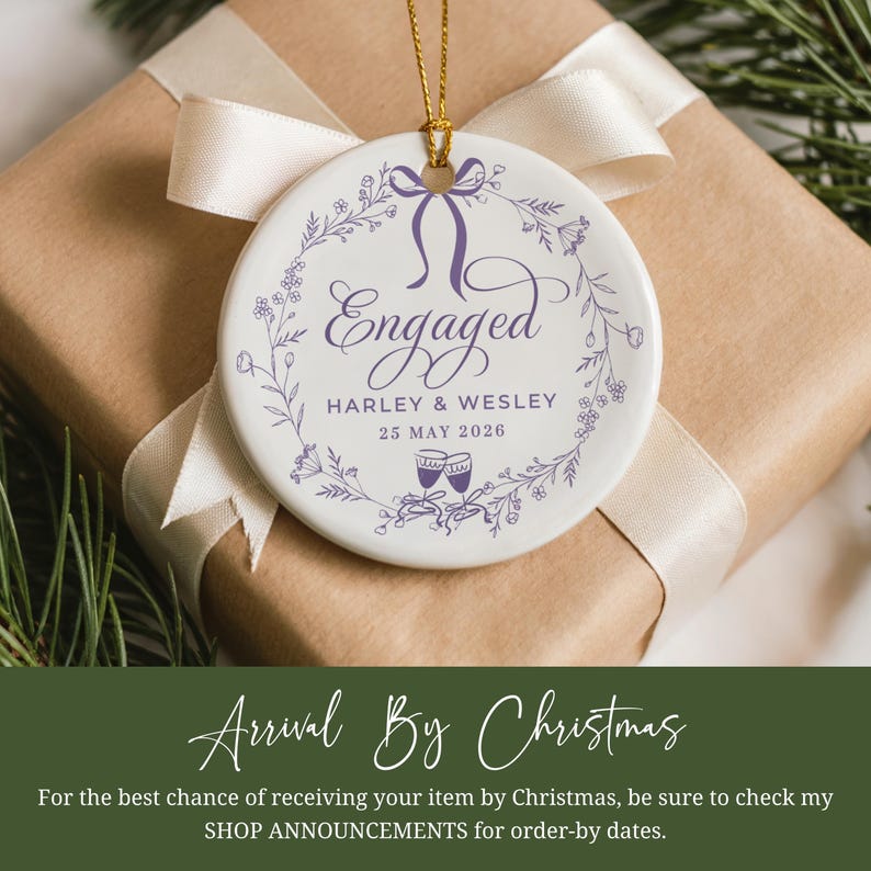 May include: A round white ceramic ornament with the word "Engaged" in purple script, encircled by a floral wreath. It features the names "Harley & Wesley" and the date "25 MAY 2026". The ornament hangs on a gold string, presented on a wrapped gift.