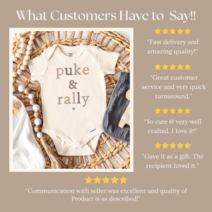 May include: A beige baby onesie with the text "puke & rally" in pink, brown, and gray lettering. The onesie is on a woven wicker surface with a gray and white fabric square and a wooden toy.
