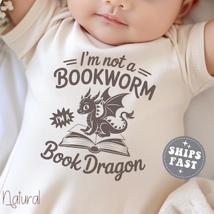 May include: A natural-colored baby onesie with the text "I'm not a BOOKWORM I'M A Book Dragon" and a graphic of a dragon sitting on a book. The onesie has short sleeves and a round neckline. The text "Natural" is printed at the bottom left.