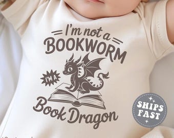 Może przedstawiać: Naturalne body dla niemowląt z napisem "I'm not a BOOKWORM I'M A Book Dragon" i grafiką smoka siedzącego na książce. Body ma krótkie rękawy i okrągły dekolt. Słowo "Natural" jest wydrukowane w lewym dolnym rogu.