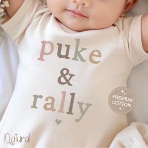 May include: A white baby onesie with the text "puke & rally" in a multi-colored font. The onesie has a small heart at the bottom and a "Premium Cotton" label.