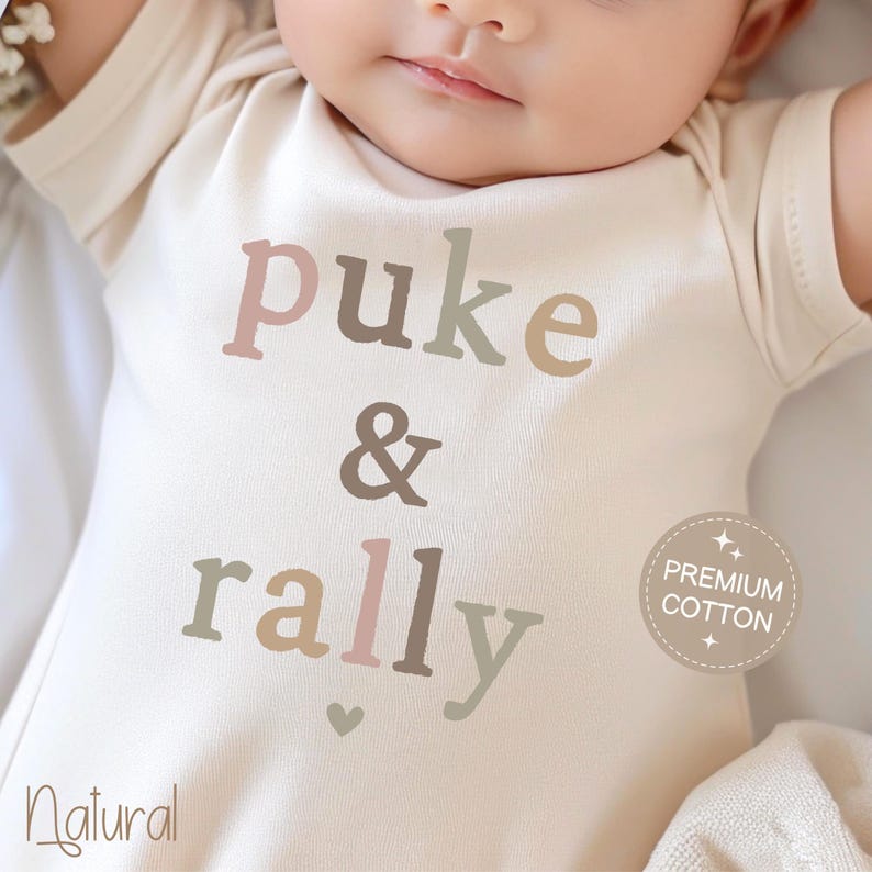 May include: A white baby onesie with the text "puke & rally" in a multi-colored font. The onesie has a small heart at the bottom and a "Premium Cotton" label.